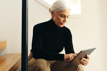 Mature business professional using a tablet for work, sitting on the stairs in her office
