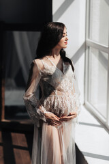 a beautiful pregnant woman in a lace transparent dress hugs her tummy with her hands near the window. Concept of pregnancy, motherhood, preparation and waiting. The beauty of a woman during pregnancy