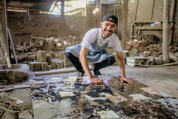 Smiling stonemason squats while washing large squares of stone in the workshop - obrazy, fototapety, plakaty