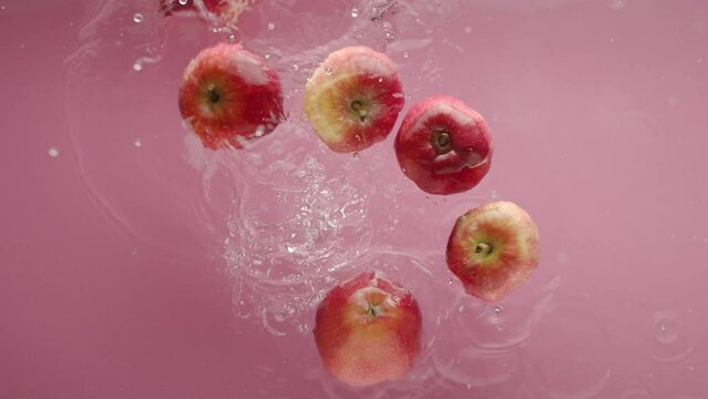 Falling apple in clear water. From above slow motion static shot of clean transparent water with pink background and falling ripe fresh apple in pile of fruit