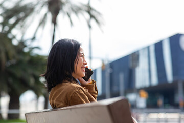 Fototapeta premium Side of young asian woman talking with cellphone while sitting on bench in city