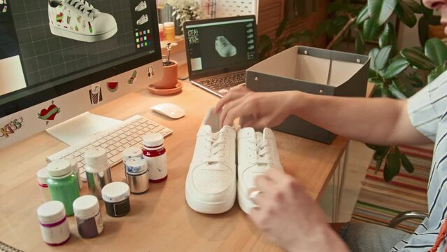 High angle medium close up of male Caucasian designer taking white sneakers out of box choosing paint to customize them