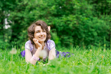 a young woman in a lilac dress lies on the green grass and smiles against the backdrop of trees in the park