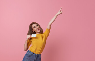 Cheerful happy woman enjoying showing credit card and hand pointing finger isolated on pink...