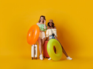 Portrait of two cheerful young women standing holding rubber ring and baggage going to travel on holidays isolated on yellow background.