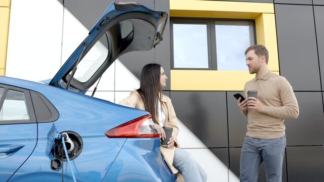 Young couple man and woman traveling together by new car having stop at charging station. Boyfriend holding cup drinking hot coffee talking to the phone and girlfriend looking aside and smiling