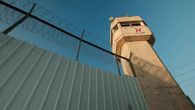 A wide angle camera panning, facing up towards a tall prison walls, with barbed wire and a watchtower at dusk, Israel.