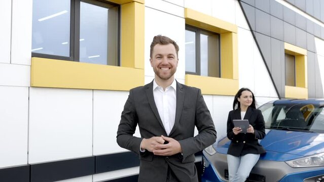 Confident male shop assistant in formal clothes holding key and looking at camera at automobile car dealer shop outdoor