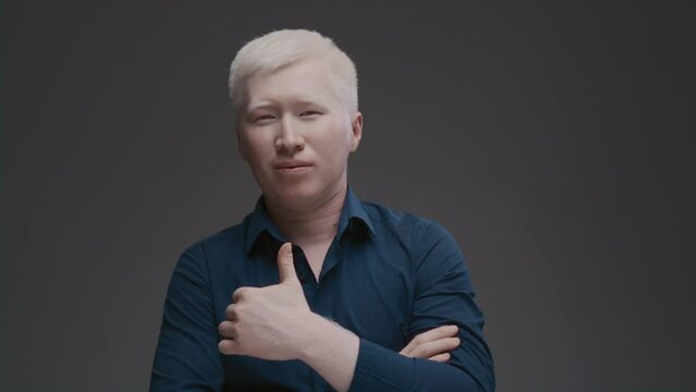 Portrait of an Asian albino guy. An eastern man with a genetic mutation of pigment shows a like sign with his hand. White hair and eyebrows. The male is wearing a dark shirt in the studio