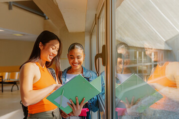 Latina girl and Asian girl, young students, happily review their class notes in the hallway of their university campus or high school. back to school