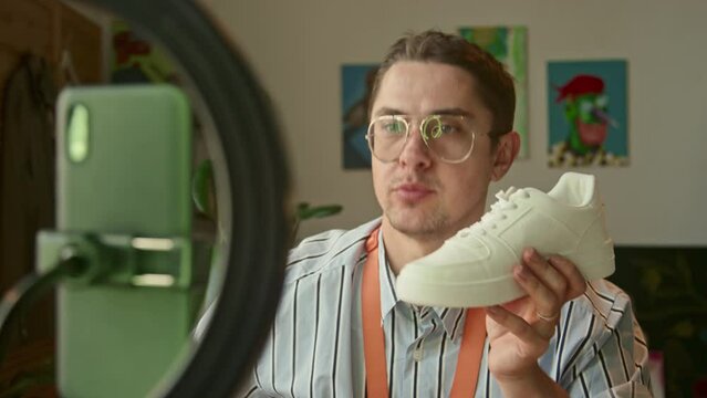 Medium close up of cheerful male designer shooting video lesson showing white sneakers at camera and having blooper in the end