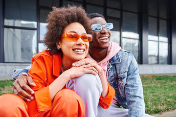 Pleased young african american woman in sunglasses and bright outfit looking away while spending time with best friend and sitting on blurred urban street, friends with trendy aesthetic