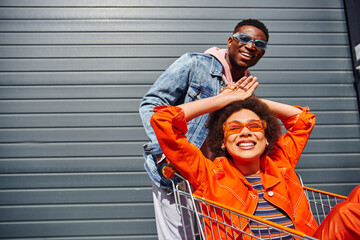 Cheerful young african american woman in sunglasses and bright outfit sitting in shopping cart near...