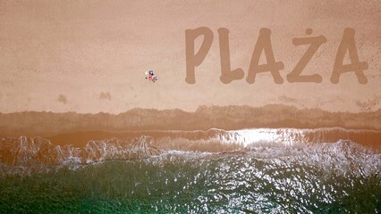 Widok z lotu ptaka na piękną piaszczystą plażę nad błękitnym morzem, gdzie leży i opala się dwóch wczasowiczów, obok nich napis 