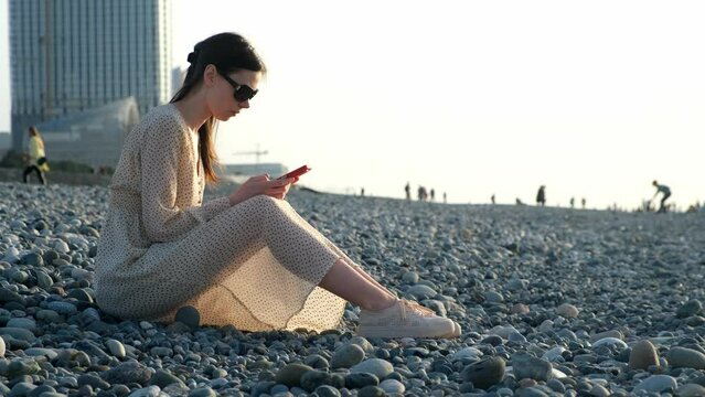 Thoughtful woman browsing smartphone sitting on pebble sea coast in Batumi city Georgia. Female chatting online in social media in phone. Summer vacation, beach resting travel tourism journey to sea.