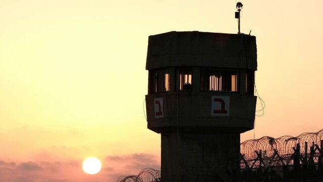 A silhouette of a prison watchtower at sunset. the sun ball is beautifully set at the bottom of the frame, while some barbed wire is apparent as well.