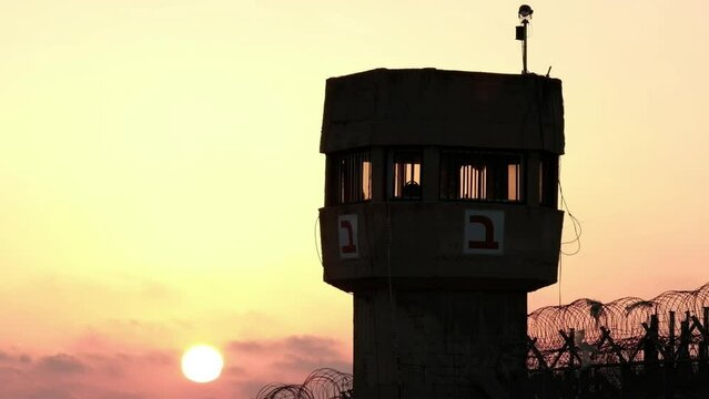 A silhouette of a prison watchtower at sunset. while the sun ball is nicely set at the bottom of the frame, a military helicopter is passing right underneath it. a 4K video clip, Israel.