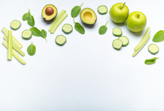 Healthy food border with green fresh vegetables and fruit on simple white background: apples, spinach, celery sticks, spinach leaves, cut avocado, cucumber. Space for text
