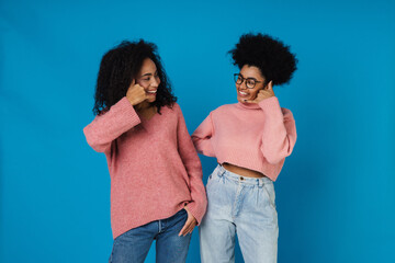 Two positive african women showing call gesture and looking at each other isolated over blue background