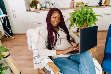 Pregnant black woman using laptop while sitting in armchair