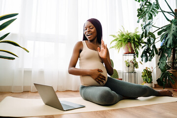 Black pregnant woman gesturing and using laptop during yoga practice