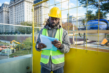 front view of young black engineer man and construction site manager reading document outdoors.
