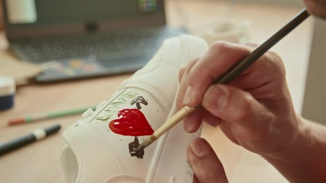 Selective close up focus on unrecognizable person holding paint brush and drawing heart picture on white sneakers indoor at daytime