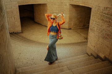 Fashionable woman wearing trendy orange linen shirt, crochet crop top, maxi denim skirt, blue sunglasses, carrying wicker tote bag. Full-length fashion portrait. Copy, empty space for text - obrazy, fototapety, plakaty