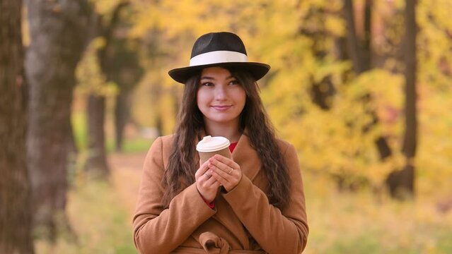 A pretty young woman in a trendy beige coat and hat walks in the park in autumn, admires the yellow leaves and drinks coffee from a disposable cup