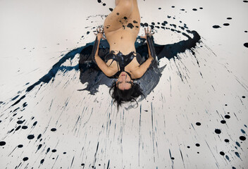 sexy naked woman partly painted in black color, lies decorative, elegant on the Studio floor, between black paint drop splashes. Creative, abstract expressive body art painting