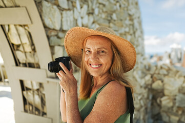 Portrait of a smiling happy elderly woman tourist traveling with a camera posing against the backdrop of mountains. Old elegant lady in straw hat on grass at countryside. Active retired people concept
