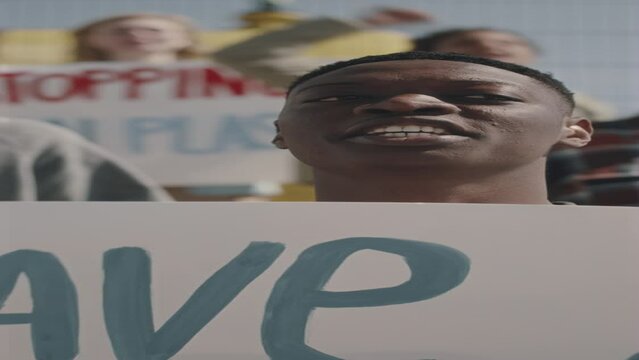 Vertical shot of young black man holding poster, looking at camera and hanting slogan while protesting with environmental activists against pollution