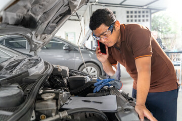 Naklejka na ściany i meble An Asian male car mechanic using his phone while working on repairing a car's engine at car repair garage