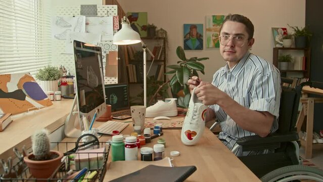 Panning right portrait of male Caucasian artist sitting in wheelchair in loft studio customizing white sneakers and looking at camera
