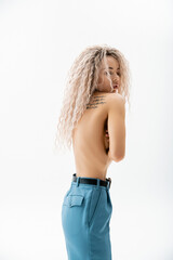 seductive and expressive woman with shirtless tattooed body obscuring naked breast with hands while standing in blue oversize pants on grey background, fashion and sexuality, modern individuality