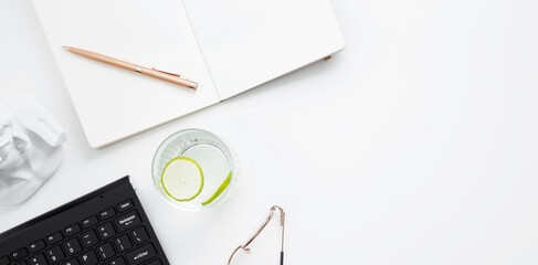 Workplace with white notebook, black keyboard, stationery, glass of water with lime and glasses on white desk. Flat lay office desk, mock up space for text. Top view. Copy space