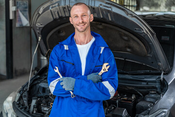 A happy male car mechanic in blue jumpsuit holding a wranch in each hands and cross his arms while standing in front of a car with hood open