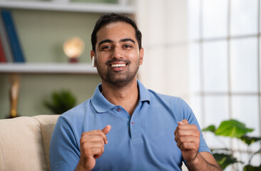 Happy indian man talking by looking camera on video call while sitting on sofa at home - concept of Virtual connection, Online interaction and Digital communication