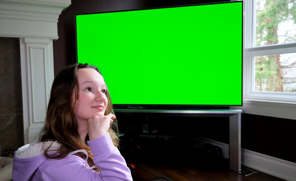 teenage girl sits admiringly in front of a big green chroma key tv monitor advertising space she enthusiastically points finger laughs thumbs up admiration 