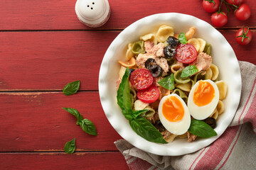 Italian pasta salad. Orecchiette pasta with tuna, tomato cherry, olive, basil and parmesan cheese in plate on red old rustic table background. Traditional Italian cuisine. Flat lay. Copy space.