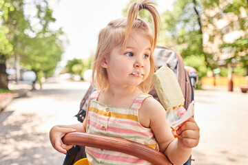 A hungry child enjoys eating ice cream outdoors. Portrait of a cute baby licking a creamy ice cream while walking in a city park, outdoor activities.