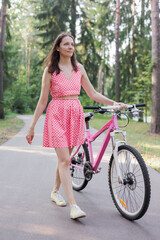 young woman rides a bike in park and walks outdoors. happy female cyclist in pink clothes is dying and actively spending weekend. lady walks and rolls bicycle with her hand