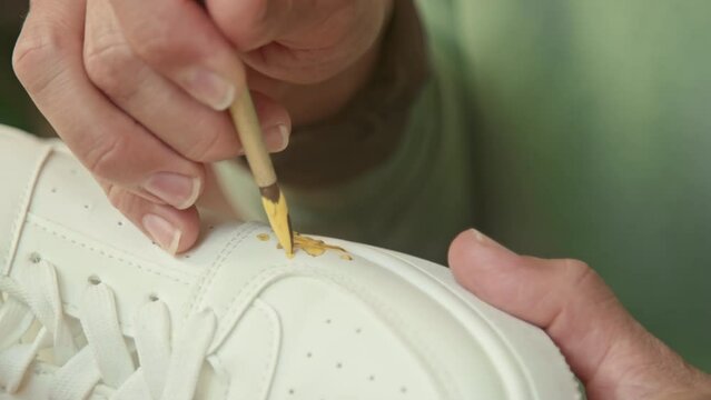 Extreme close up of hands of unrecognizable person holding paint brush and drawing yellow star on white sneaker indoor at daytime