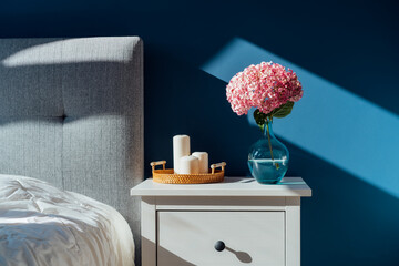 Stylish modern bedroom in dark colors. Cozy interior with navy blue walls, home decor. Bed with grey fabric headboard, white blanket, bedside table, vase with pink hydrangea flower, candles on tray.