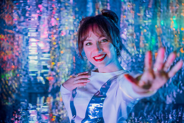 Happy smiling woman in white clothes dancing in neon light and extends a hand inviting you. Music lover. White dress code party, disco. Enjoy moment on shiny background. Selective focus