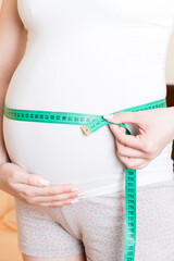 Young beautiful pregnant girl measuring her belly with a tape in home on bed. Healthy pregnancy concept