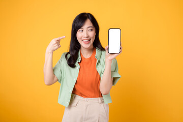 Portrait beautiful young asian woman happy smile dressed in orange shirt showing pointing finger hand gesture to smartphone isolated on yellow studio background. app smartphone concept