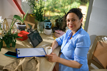Pregnant woman floral designer taking sip of coffee, smiles looking at camera while a coffee break...