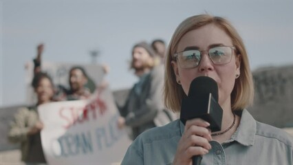 Female TV reporter looking at camera and talking in mic while reporting on protest against environmental pollution, group of activists with posters in background