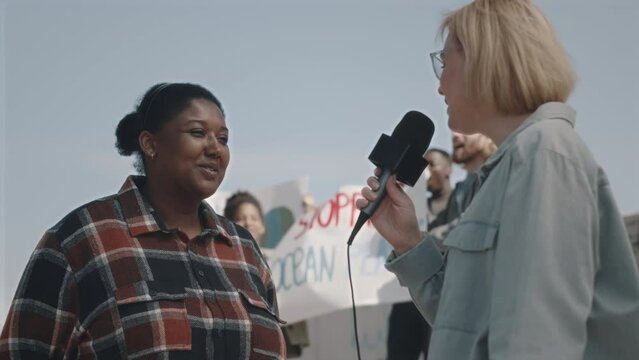 Female TV reporter holding mic and interviewing African American environmental activist during protest against pollution